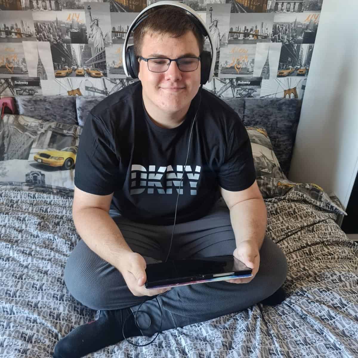 A person in cozy loungewear from DKNY sits cross-legged on a bed, headphones over their glasses, holding a tablet. A city-themed wallpaper sets the urban backdrop.