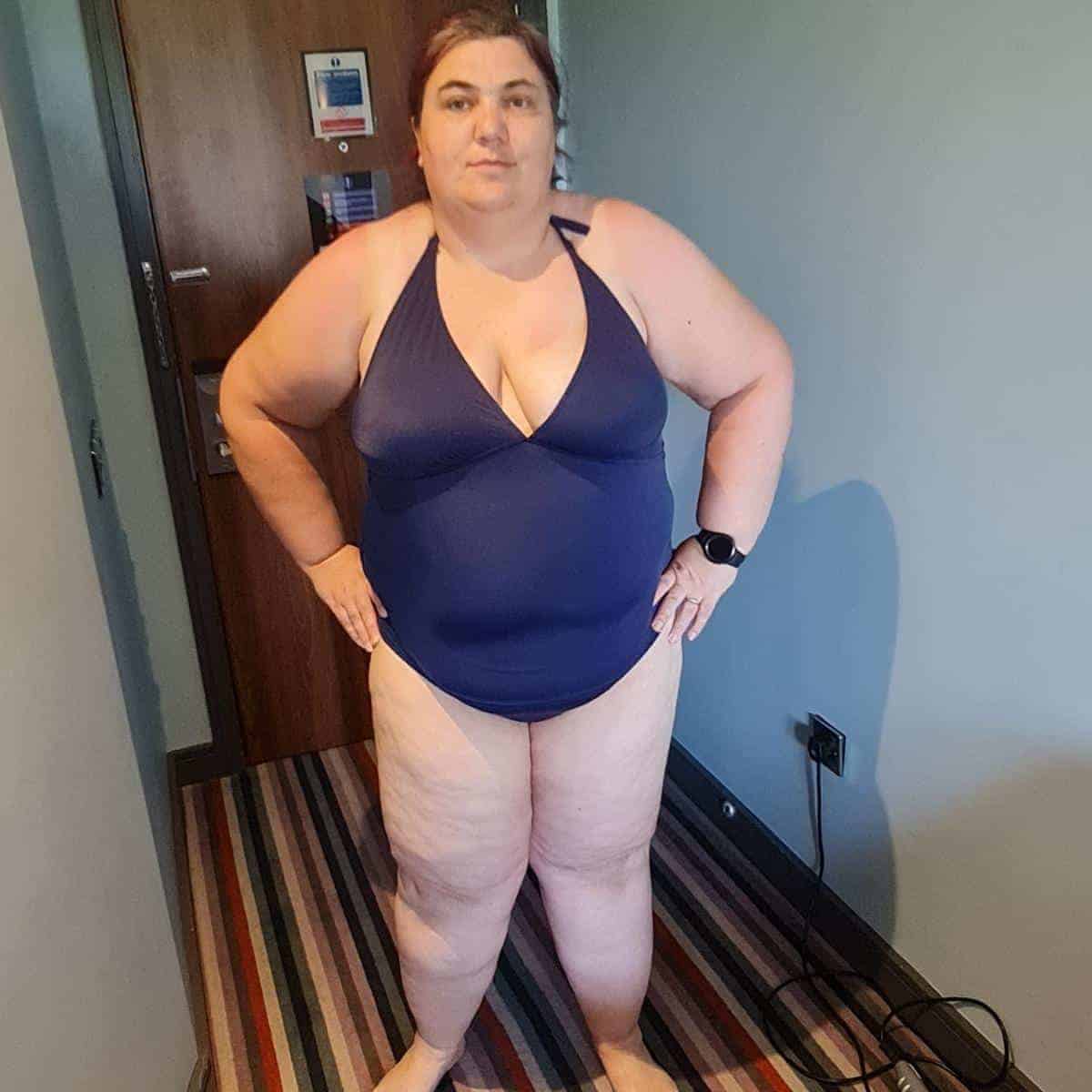 A person exudes body confidence in a blue swimsuit, standing on a striped carpet in the room, with hands confidently placed on hips.