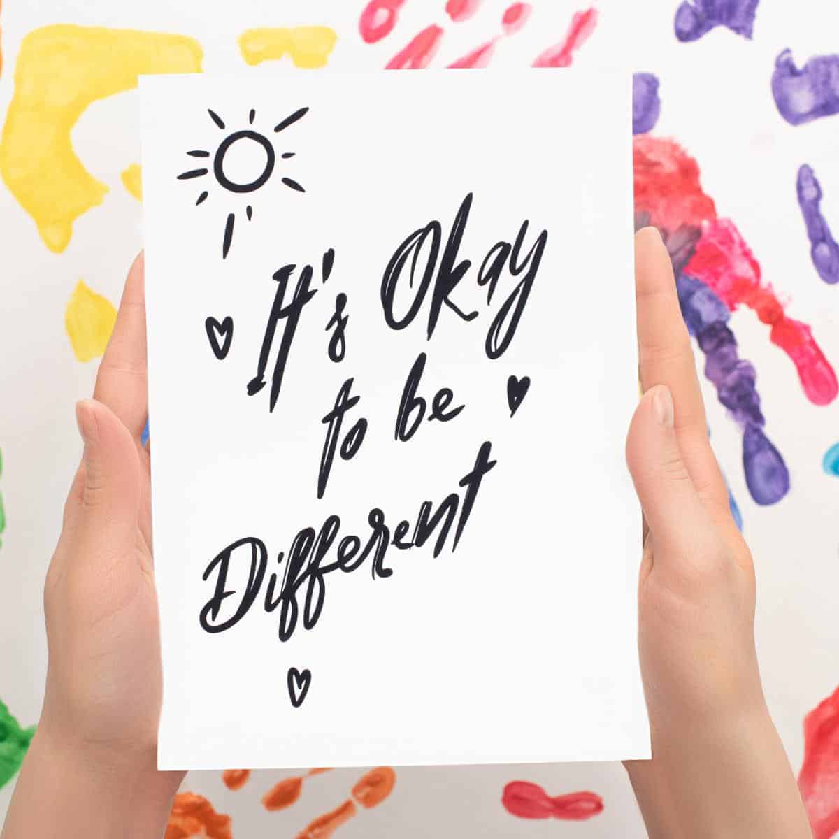 Its ok to be different written on paper held in hands with paint in background