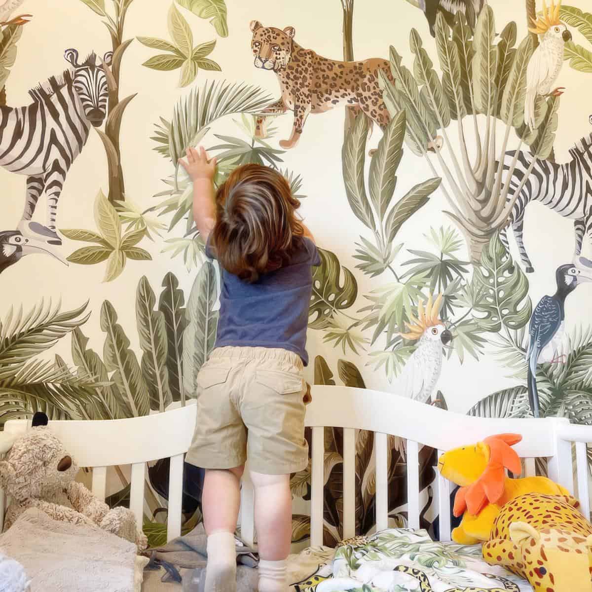 Child reaches up while standing on a bed against a jungle-themed wallpaper mural featuring animals like a zebra, leopard, and birds. Stuffed animals and a dinosaur toy are scattered on the bed.