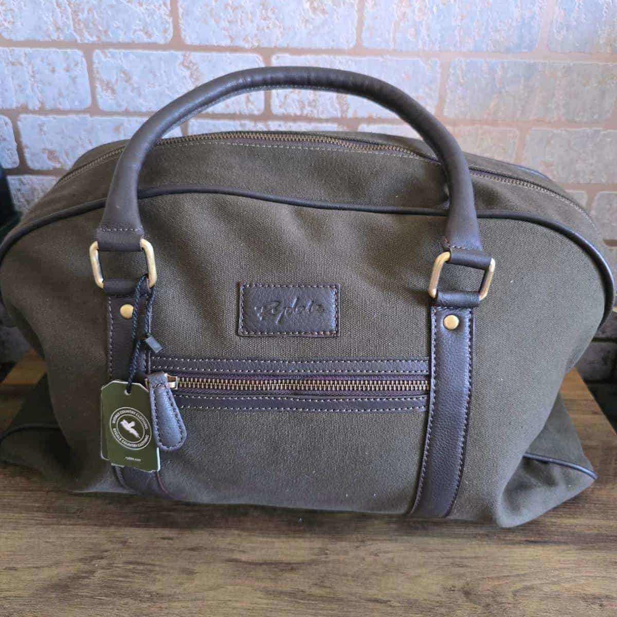 A green canvas duffel bag with dark brown leather handles and trim sits on a wooden surface. The bag has a front zipper pocket, gold metal hardware, and a stitched logo patch. A green tag is attached to one handle. Perfect for gift ideas for dad who has everything!