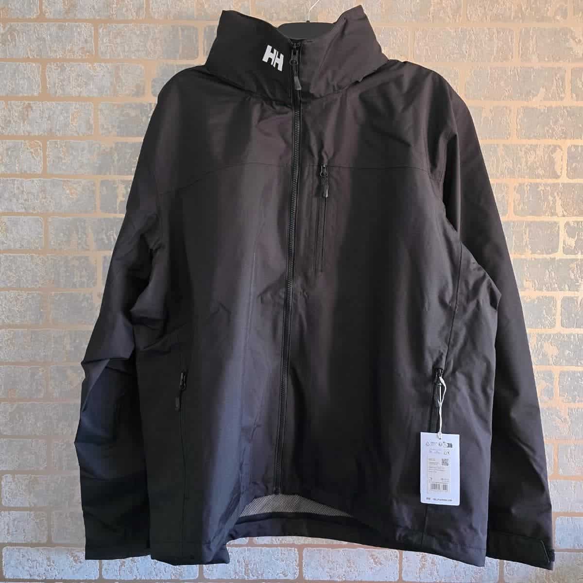 A black Helly Hansen jacket hangs against a light brick wall. The jacket has a high collar with the "HH" logo in white, zippered chest and side pockets, and a visible tag attached to the right pocket. The jacket is new and unworn.