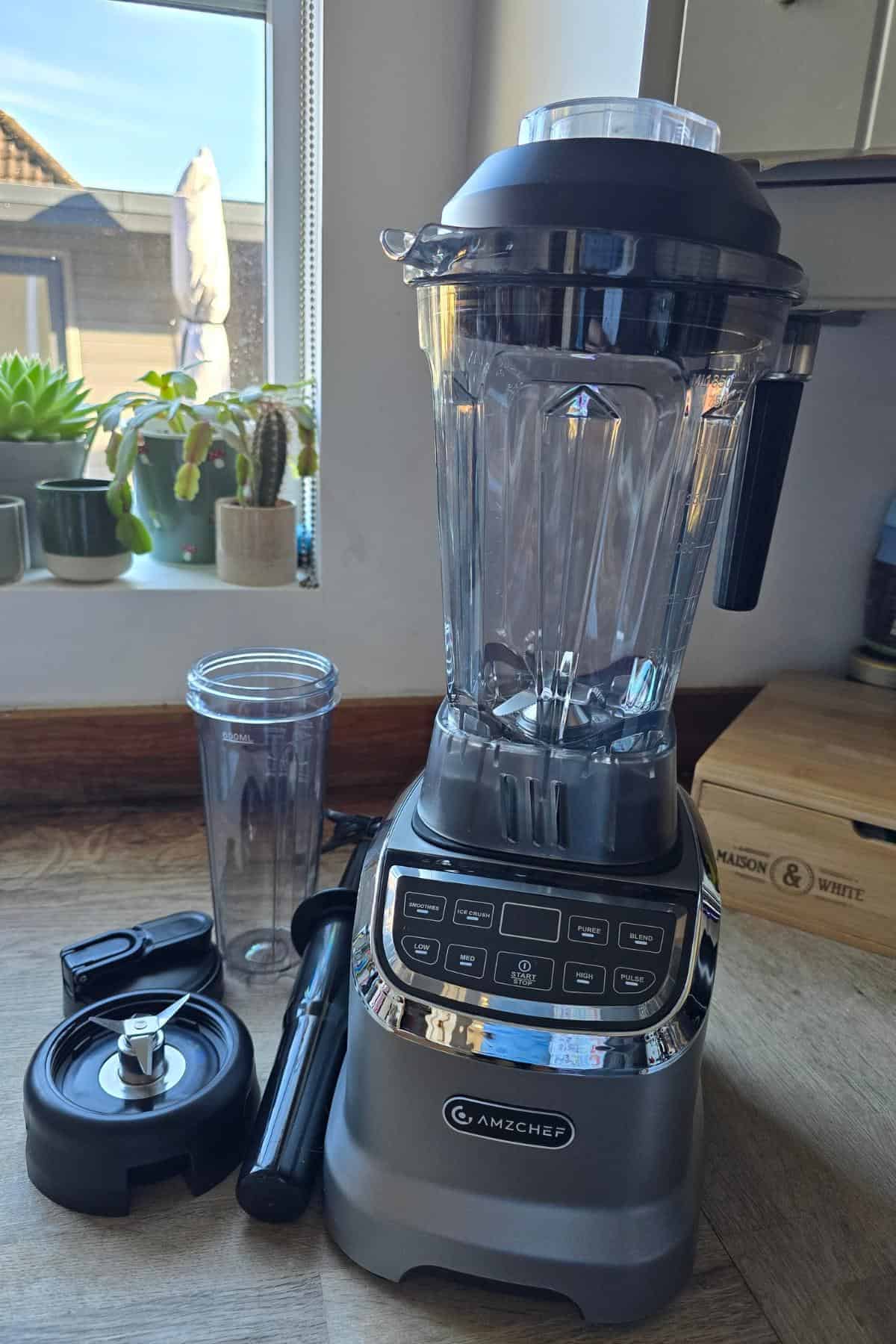 An AMZCHEF blender with a clear plastic jug, black lid, and control panel sits on the kitchen counter. Nearby are a tall blending cup, black tamper tool, blade attachment, and wooden box. Plants line the windowsill behind.