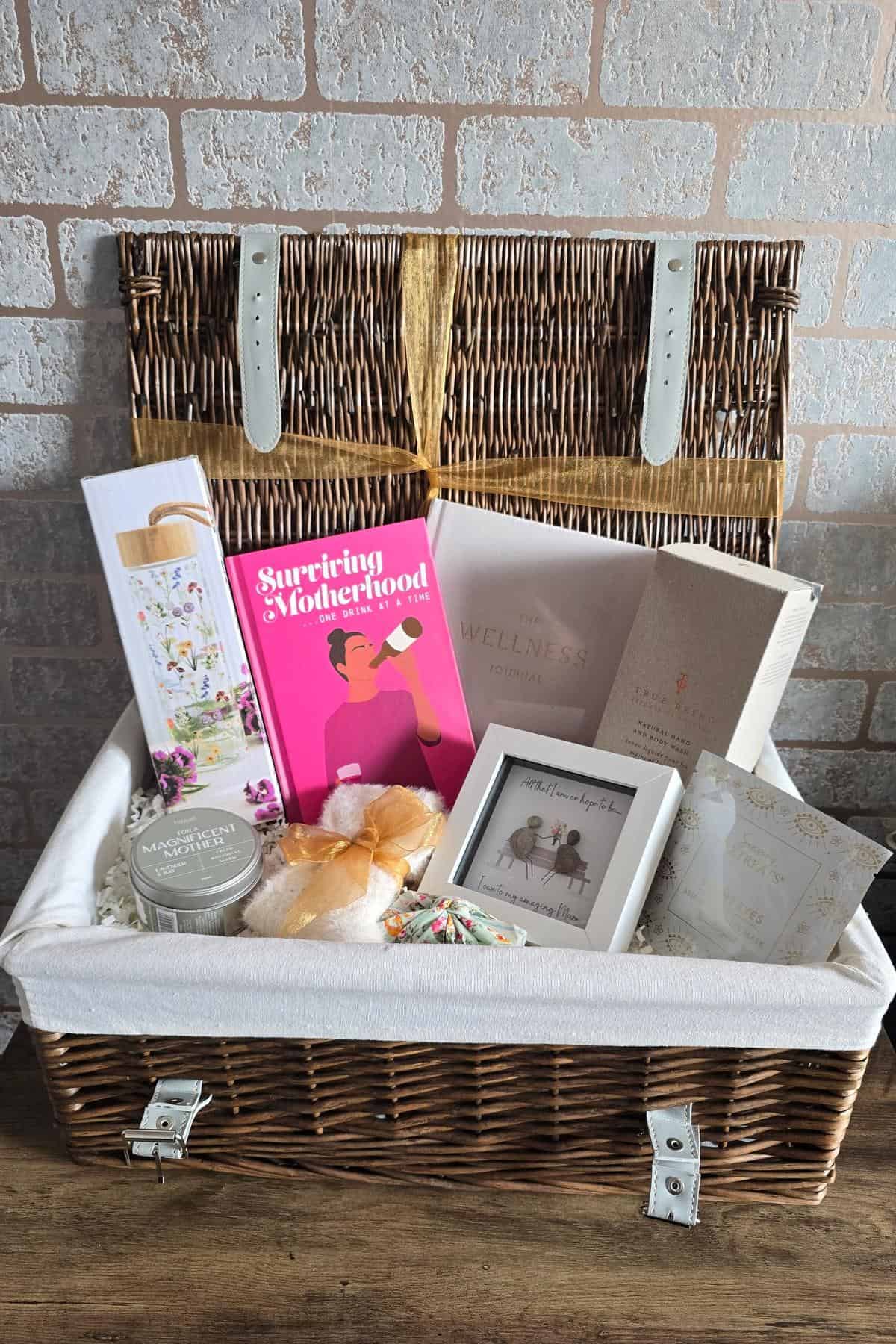 A wicker Mother’s Day gift basket sits on a wooden table against a brick wall. Inside are a pink "Surviving Motherhood" book, greeting cards, a candle, bath salts, photo frame, and soap, all arranged in a cream fabric-lined basket.