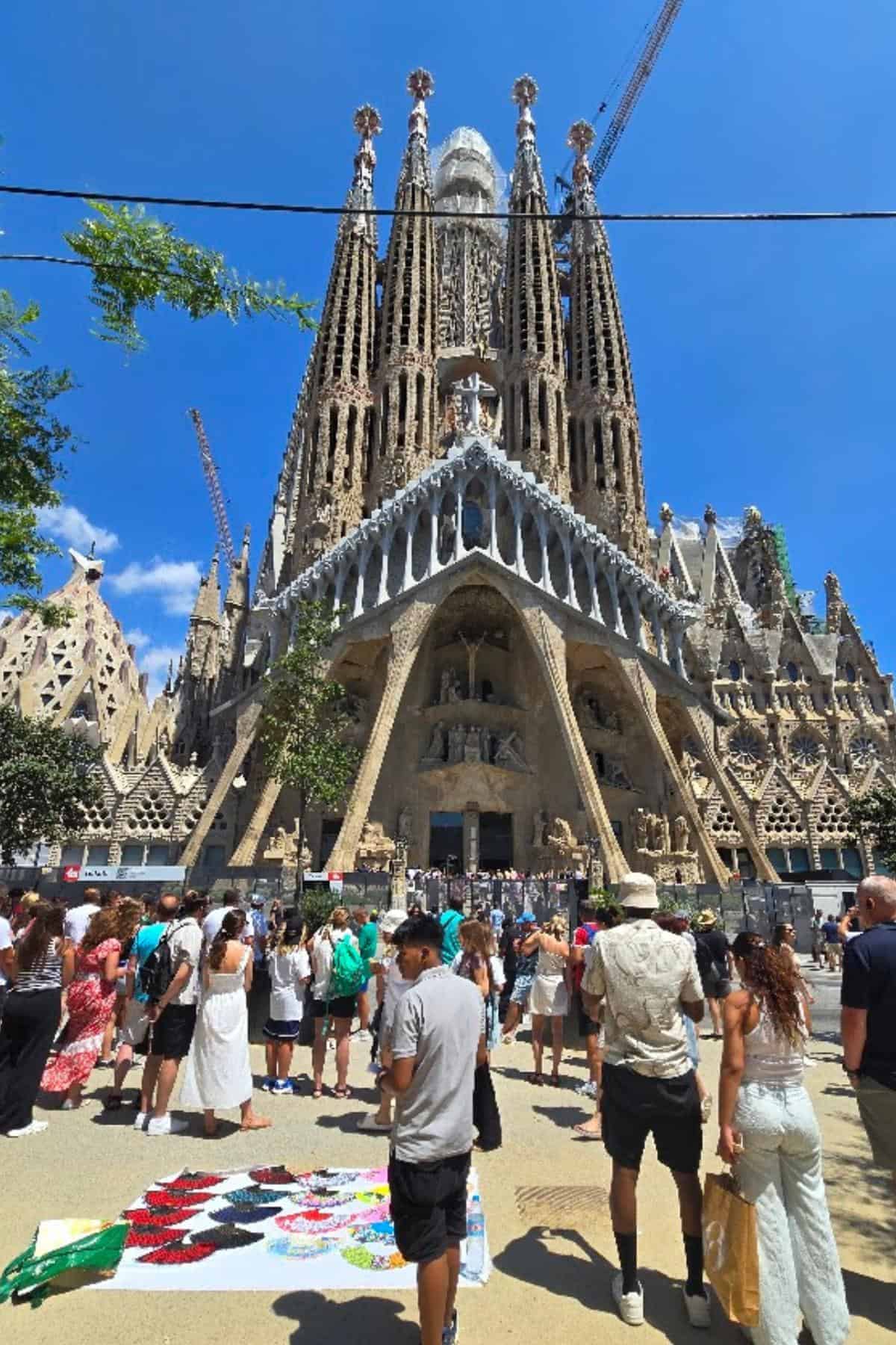 Families gather in front of the Sagrada Família basilica in Barcelona under a blue sky, with street vendors selling colorful items nearby. Tall, detailed towers and cranes rise above the cathedral, while a City Tour Bus passes in the background.