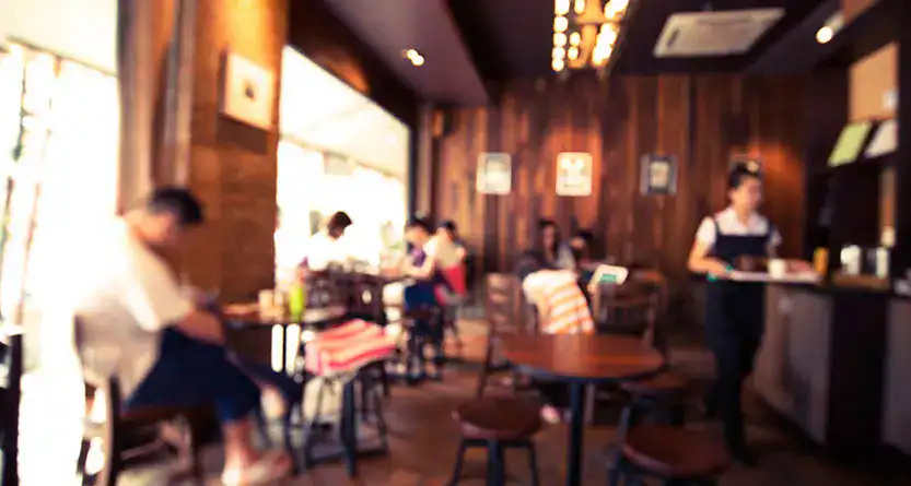 Soft-focus interior of a busy coffee shop with warm lighting—staff and customers in a café setting where branded tees and aprons build recognition.