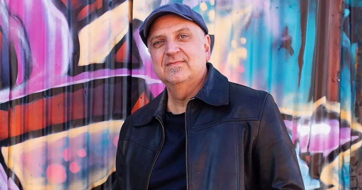 Older man in black leather jacket standing in front of colorful graffiti wall, urban street style, confident expression, music industry professional, artist, creative, street art atmosphere, hypebot content.
