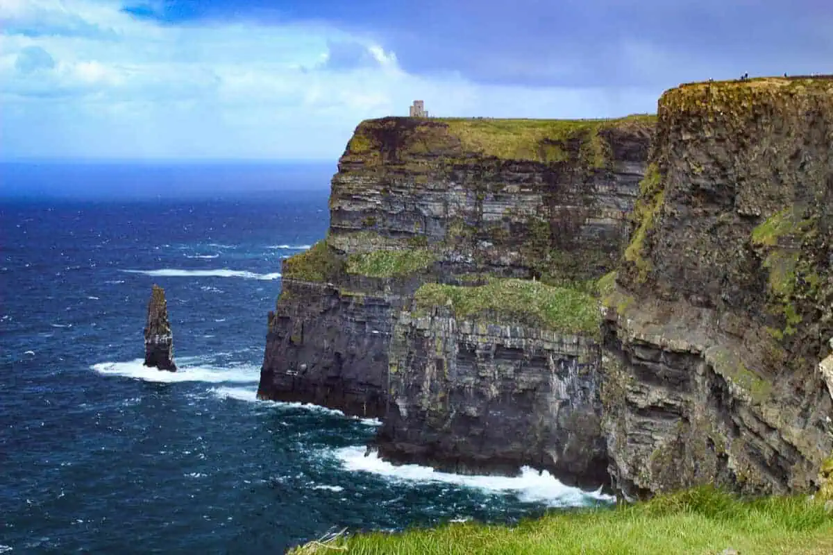 The photo showcases the majestic Cliffs of Moher on the west coast of Ireland, with the Atlantic Ocean churning below. A solitary sea stack stands near the cliff base, separated from the mainland by centuries of erosion. The cliffs rise steeply from the ocean, their stratified layers of rock visible, topped with a lush green surface. Atop the cliffs, a small stone tower or building offers a lookout point for visitors, dwarfed by the scale of the natural landscape. The sky above is a mix of blue with wispy white clouds, indicating variable weather conditions. The powerful waves hitting the cliff base create white foam, highlighting the wild and natural beauty of this Irish landmark.