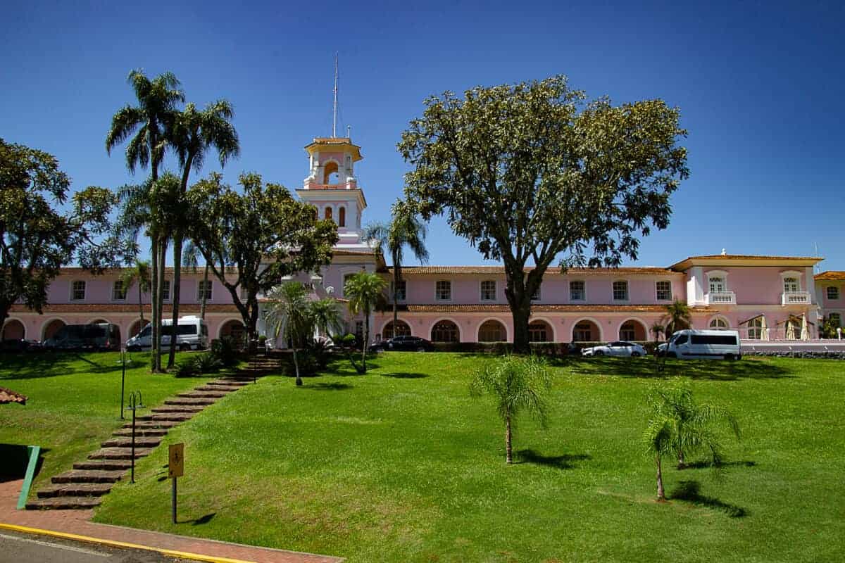 The photo showcases a picturesque two-story building painted in a soft pink hue with white trim, featuring a prominent clock tower with a spire at the center. The architecture suggests a colonial style with arched porticoes lining the front of the building. Lush, well-manicured lawns surround the structure, dotted with tropical palm trees and a large, shady tree at the forefront. The clear blue sky suggests a bright, sunny day. Stone steps lead up to the entrance, and a few vehicles are parked on the side, indicating that this may be a place of interest, possibly a hotel or historic site, which is frequented by visitors.