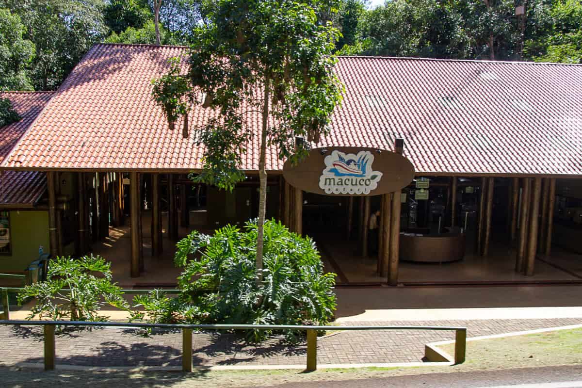 The photo shows a single-story building with a terracotta-tiled roof nestled among verdant greenery. A sign with the word "Macuco" and an illustration of a bird and leaves hangs prominently over the entrance, suggesting the building is a stop or attraction, possibly named Macuco, within a larger park or tour area. The structure has an open, pavilion-like design with large supporting wooden pillars, offering a welcoming area for visitors. The setting appears tranquil and shaded, a pleasant stop for those exploring the surrounding natural attractions. A clear sky and bright sunlight indicate a fine weather day, perfect for outdoor activities.