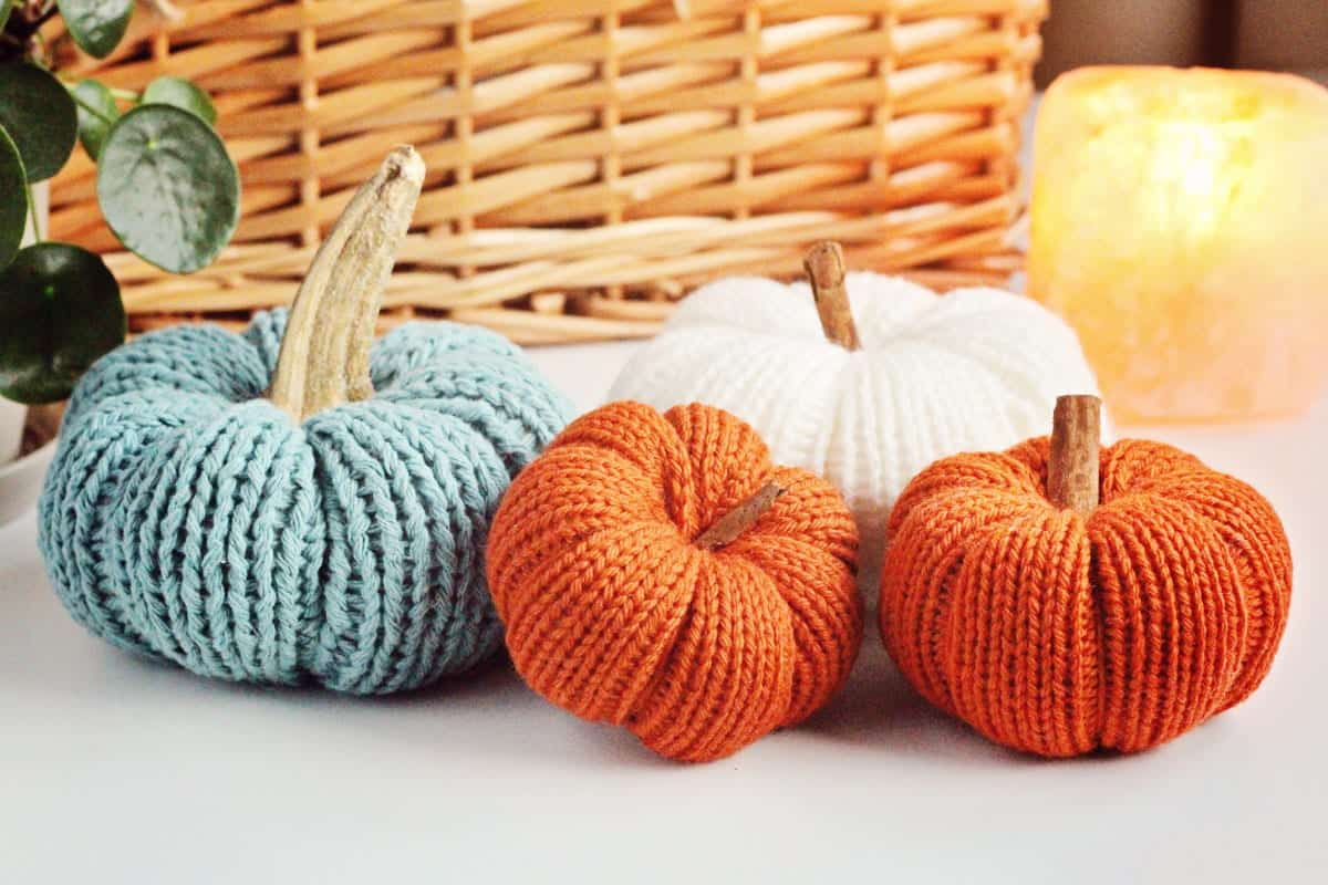 Four knitted pumpkins sitting on a white table in front of a wicker basket with a candle burning in the background. Learn how to make your own cozy pumpkin with this free knitted pumpkin pattern