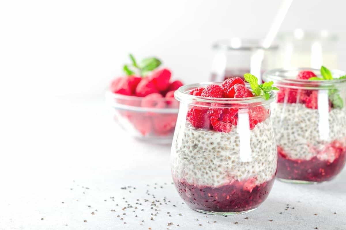 Two jars of creamy chia pudding with raspberry compote, elegantly topped with fresh raspberries and mint leaves. Great for high protein snacks or desserts.