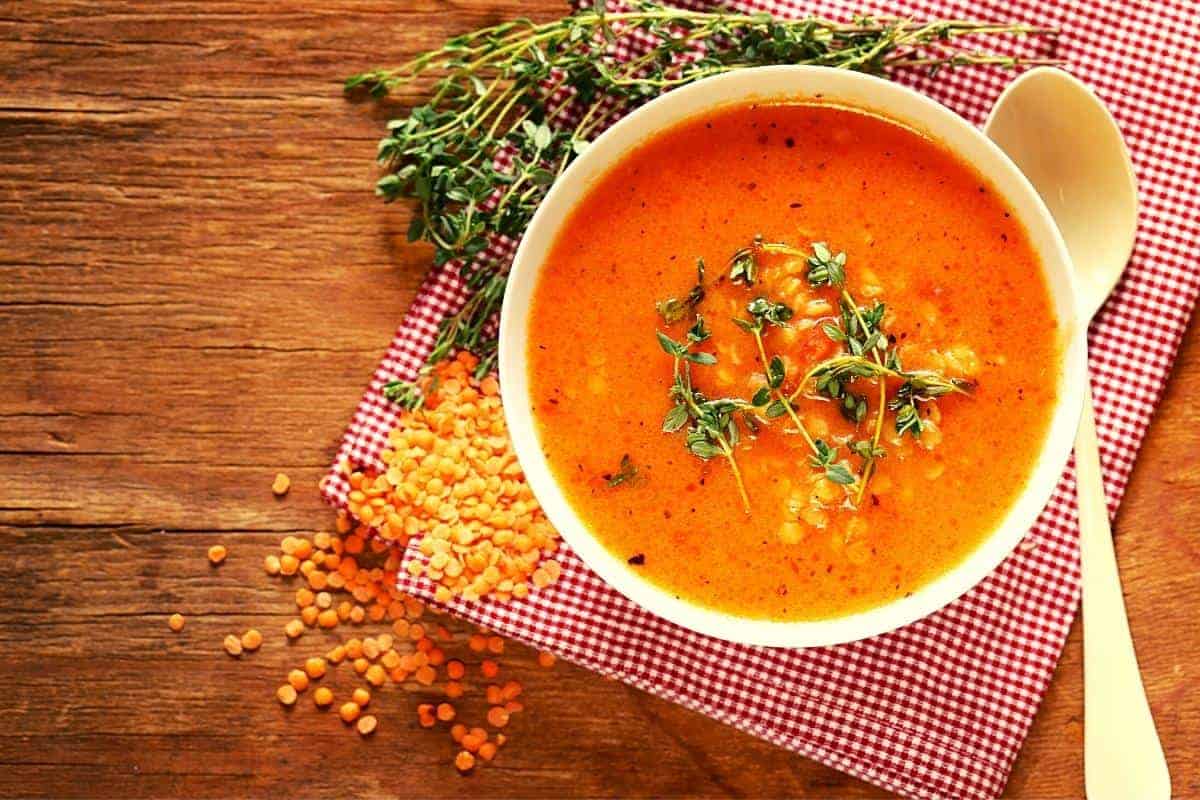 An orange coloured Lentil soup using red lentils in a bowl with some dried lentils next to the bowl.