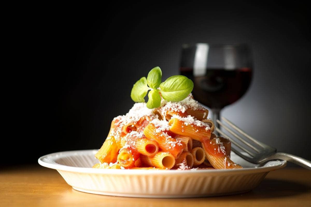 A plate of rigatoni with tomato sauce, grated cheese, and a basil garnish sits enticingly next to a glass of red wine on a rustic wooden table. This delightful scene showcases the essence of Italian foods and drink.