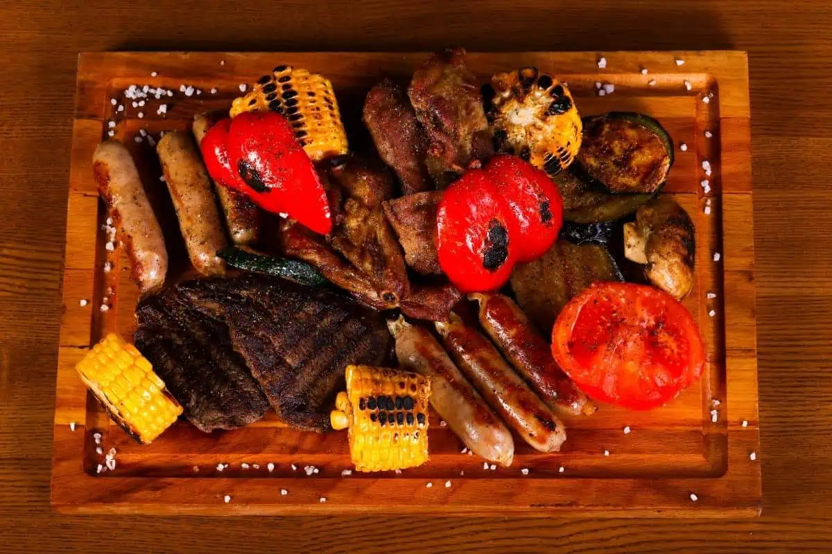 A wooden board filled with perfectly grilled meats, sausages, corn, red peppers, and zucchini slices ensures a delicious feast while taking care to avoid food poisoning.
