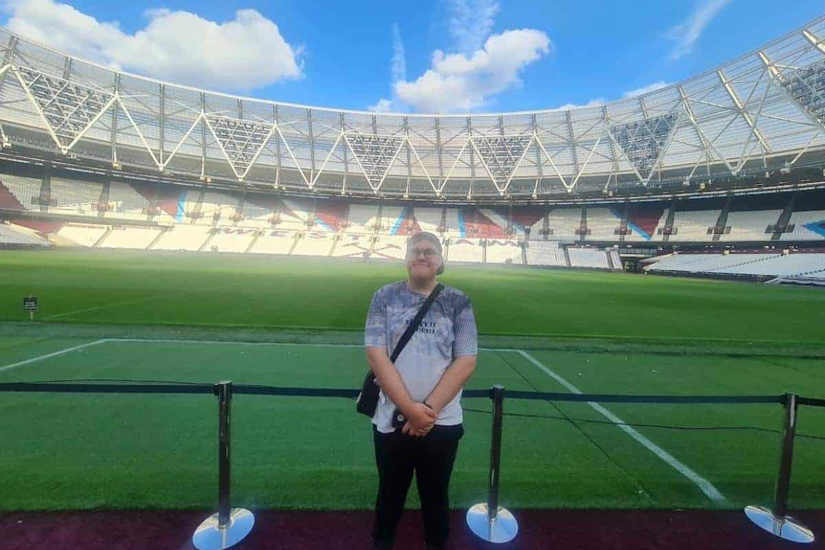 Ben, a teenage boy at the West Ham stadium tour with the pitch and seating behind him, he has a huge smile and is loving being there.
