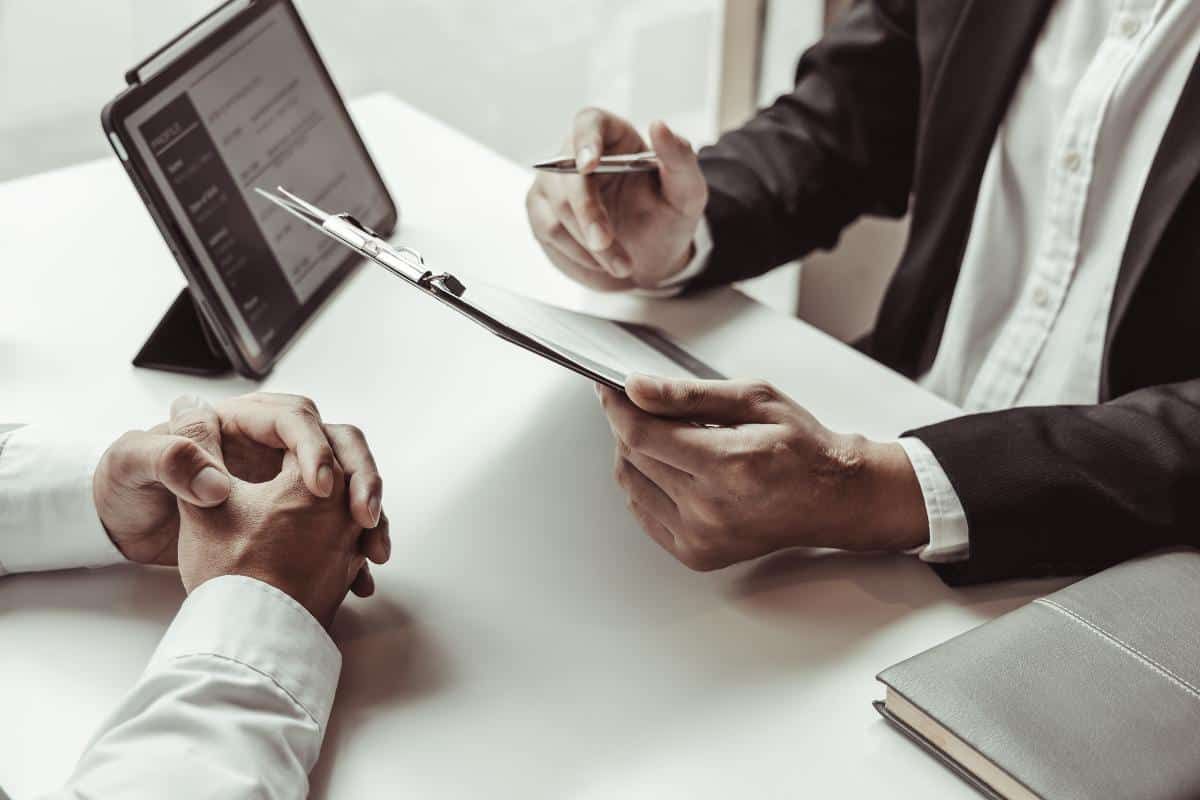 Suited man with a tablet computer on the desk holding a clipboard whilst facing another man at a desk.