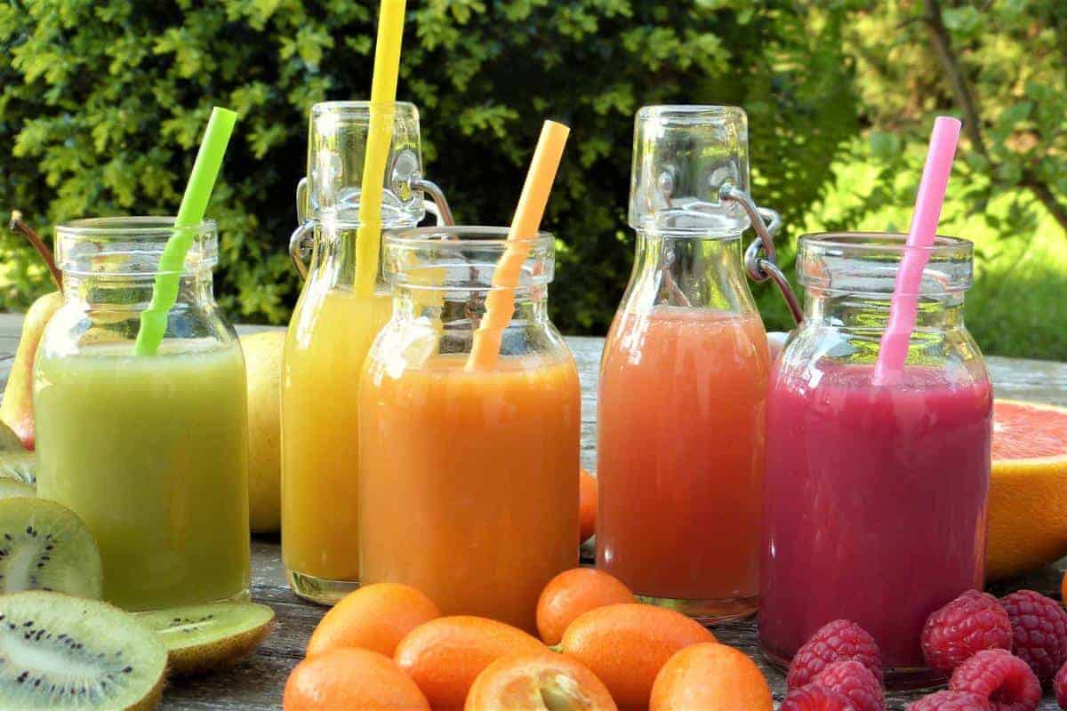 Selection of glass berrles with different colour fruit juices in and in front of the bottles sit some oranges, raspberries and kiwi.