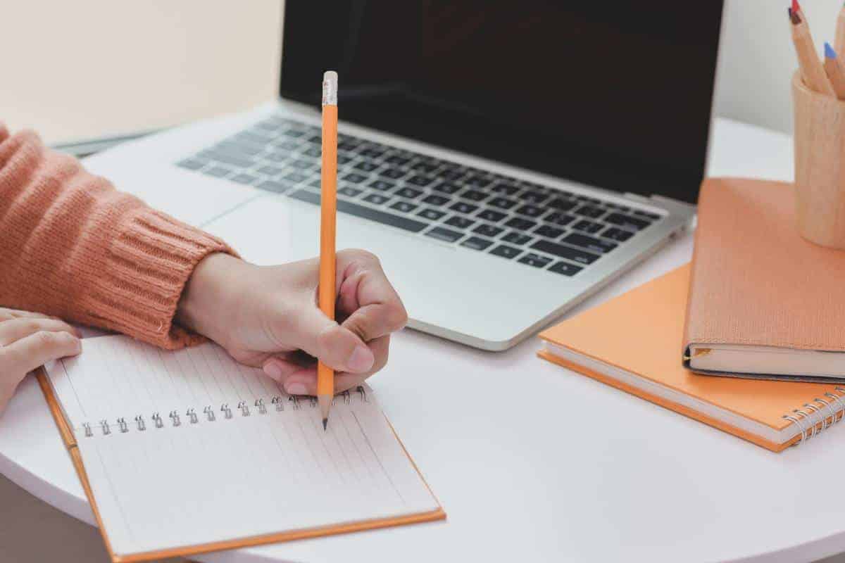 Hand writing in a notebook with a laptop in the background.
