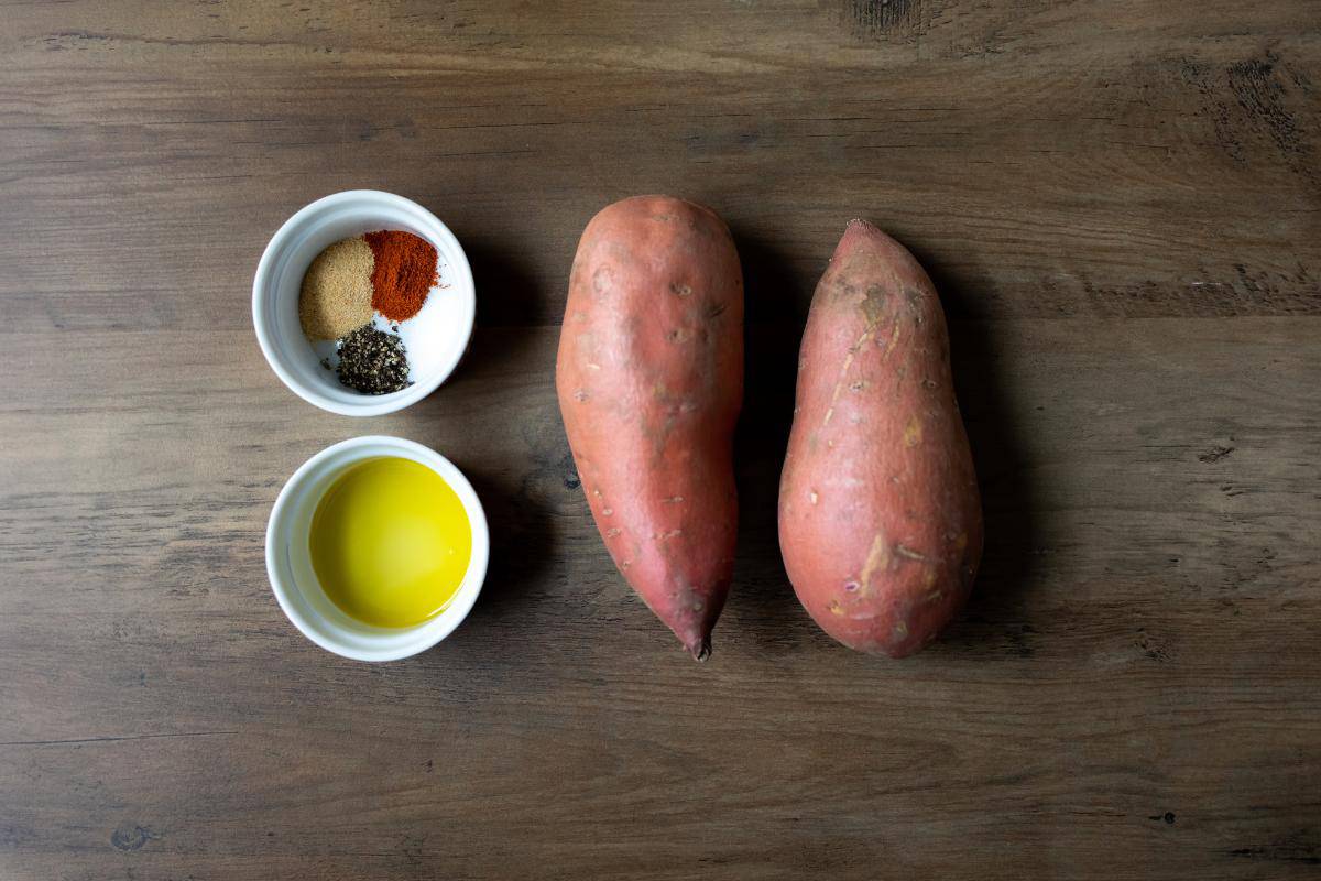 Two sweet potatoes, a small bowl with a selection of herbs and spices and another small bowl with a small amount of oil all on a wooden table.