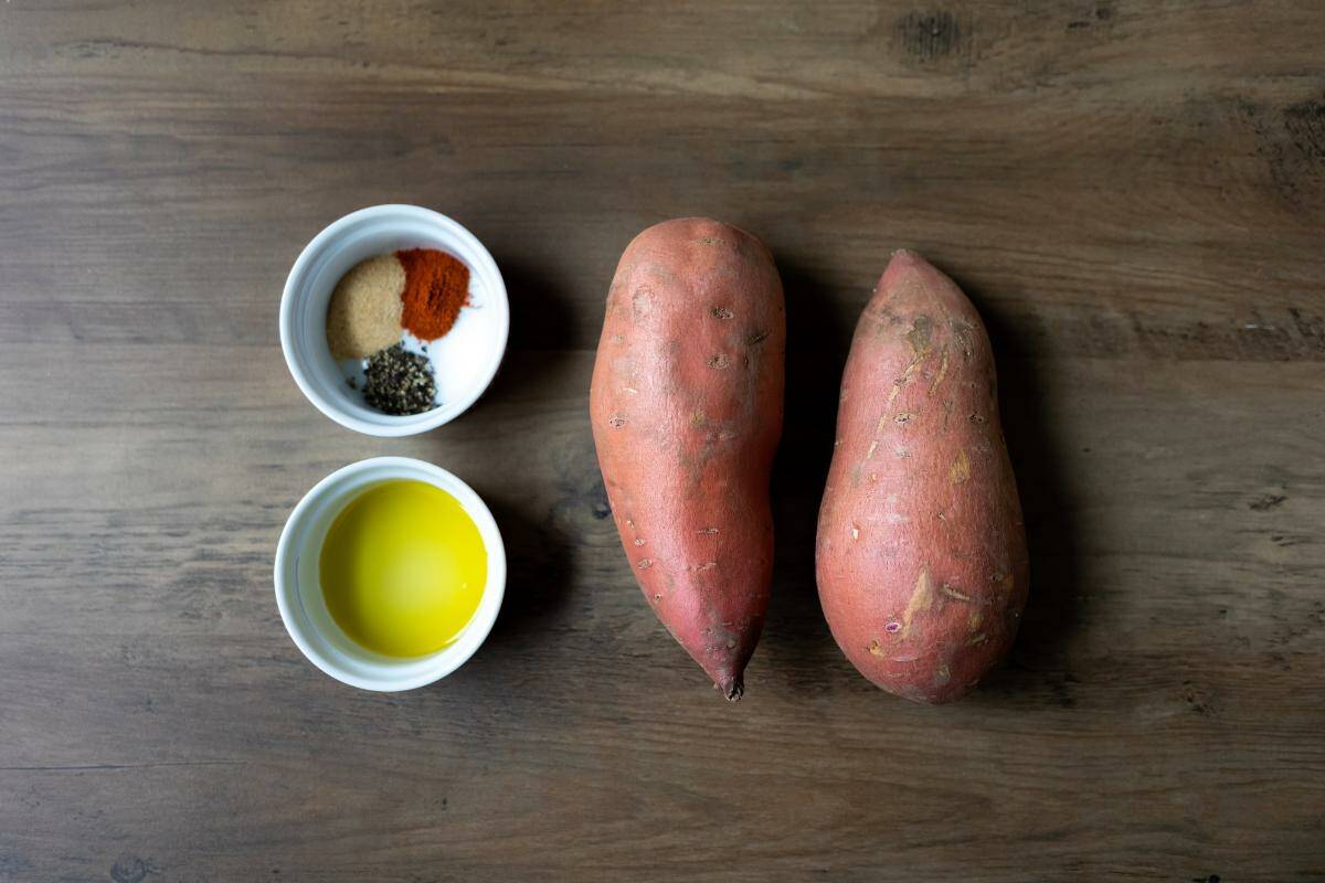 Two sweet potatoes, a small bowl with a selection of herbs and spices and another small bowl with a small amount of oil all on a wooden table.
