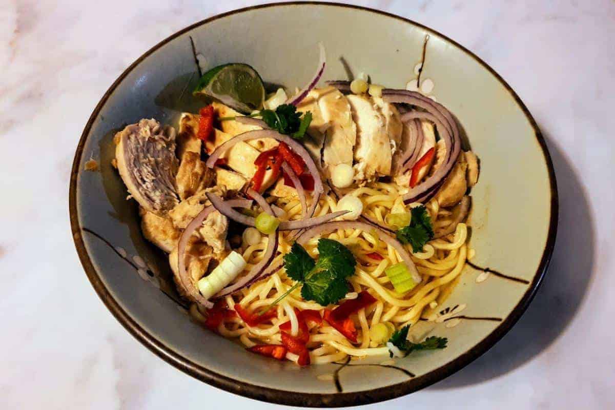 Bowl of chilli chicken ramen with chicken, red onion, spring onions and lime.