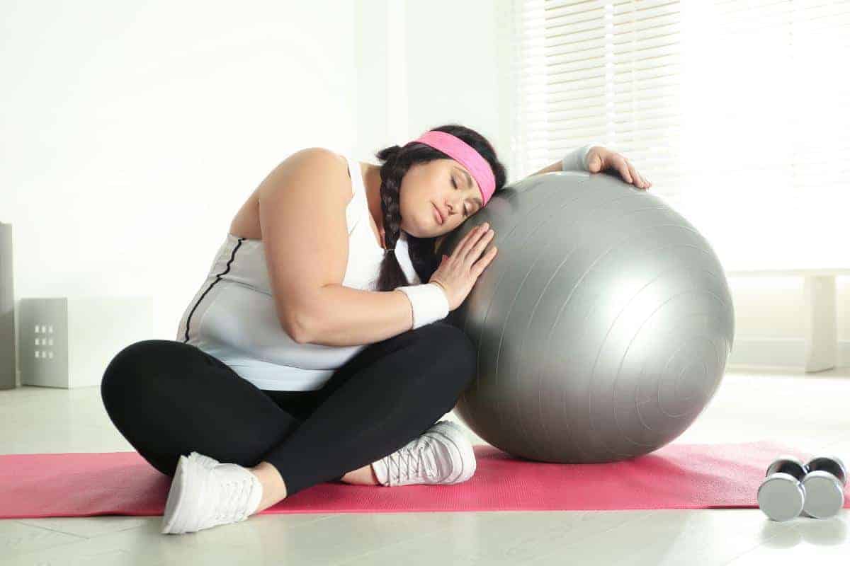 Overweight lady in fitness clothing next to a gym ball sitting on the floor.