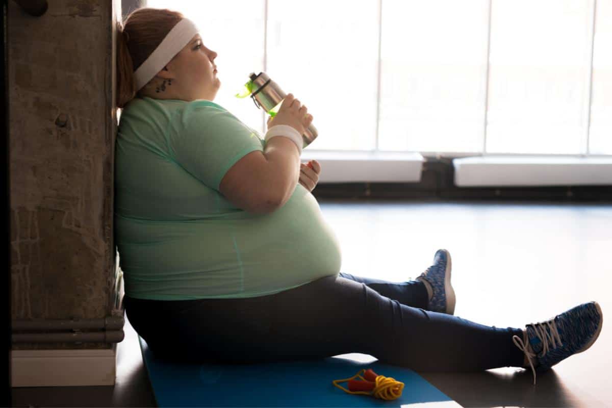 Overweight lady sitting down on exercise mat having a drink of water after exercising.