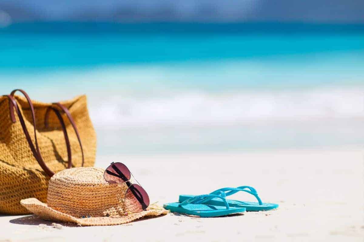 flip flops and sun hat with sunglasses on sand with blue sea in background.
