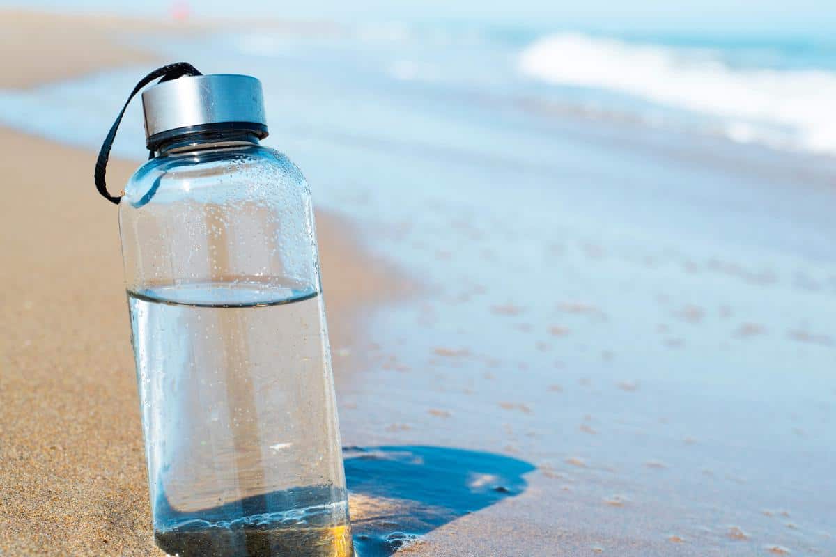 clear water bottle containing water in the sand with the beach water flowing slightly up to the bottle