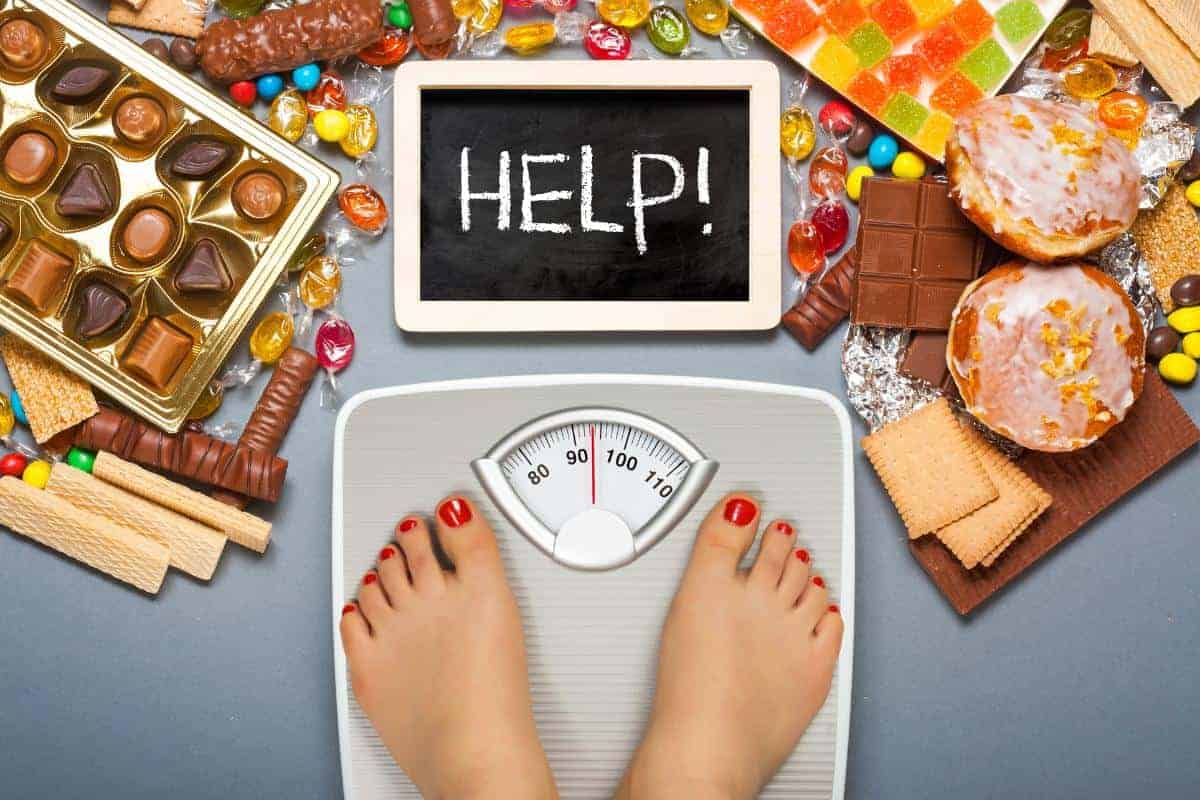 A ladies feet with red painted toenails on bathroom scales with a blackboard saying the word help and surrounded by chocolates, doughnuts and sweets.