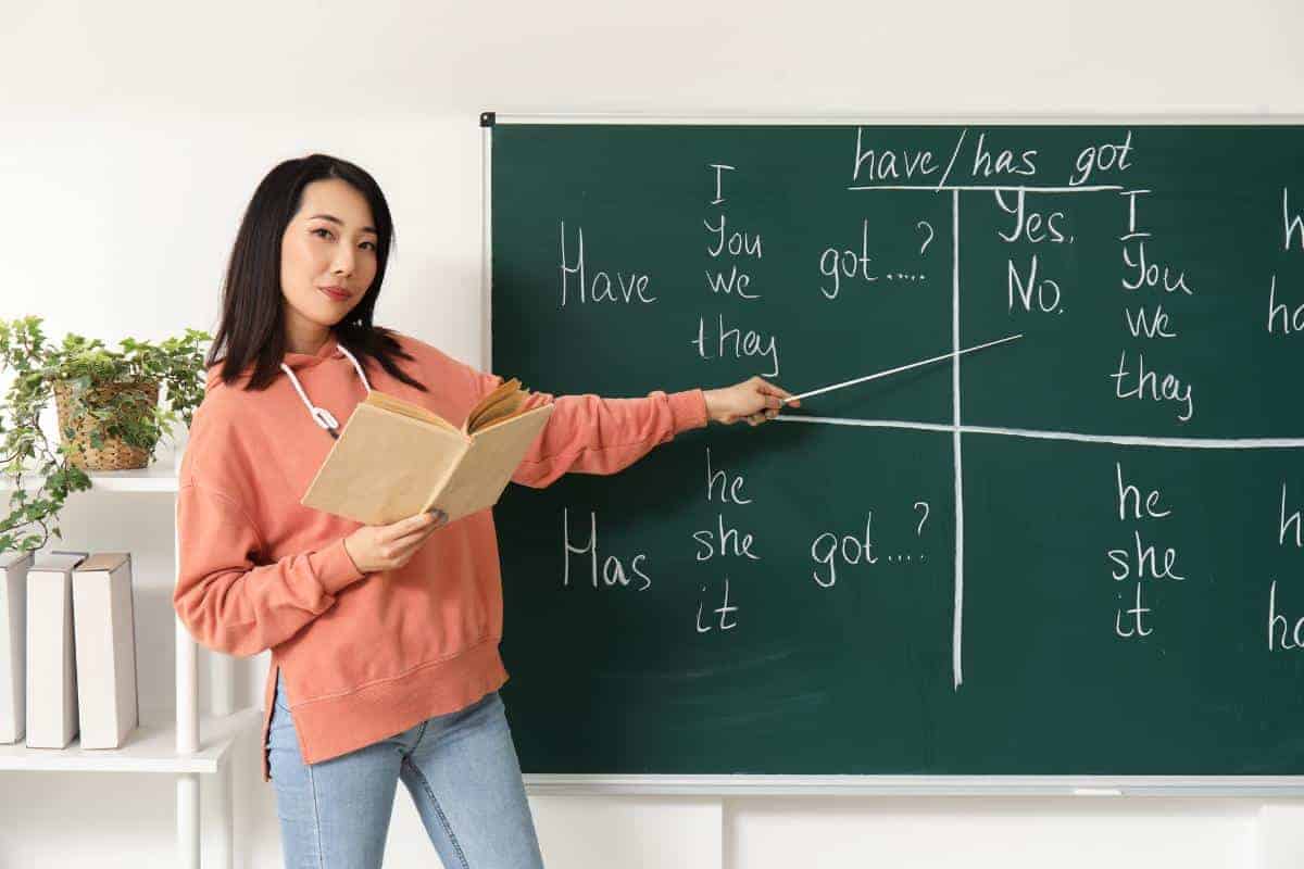 A Japanese tutor pointing to a chalk board with an English lesson written on it.