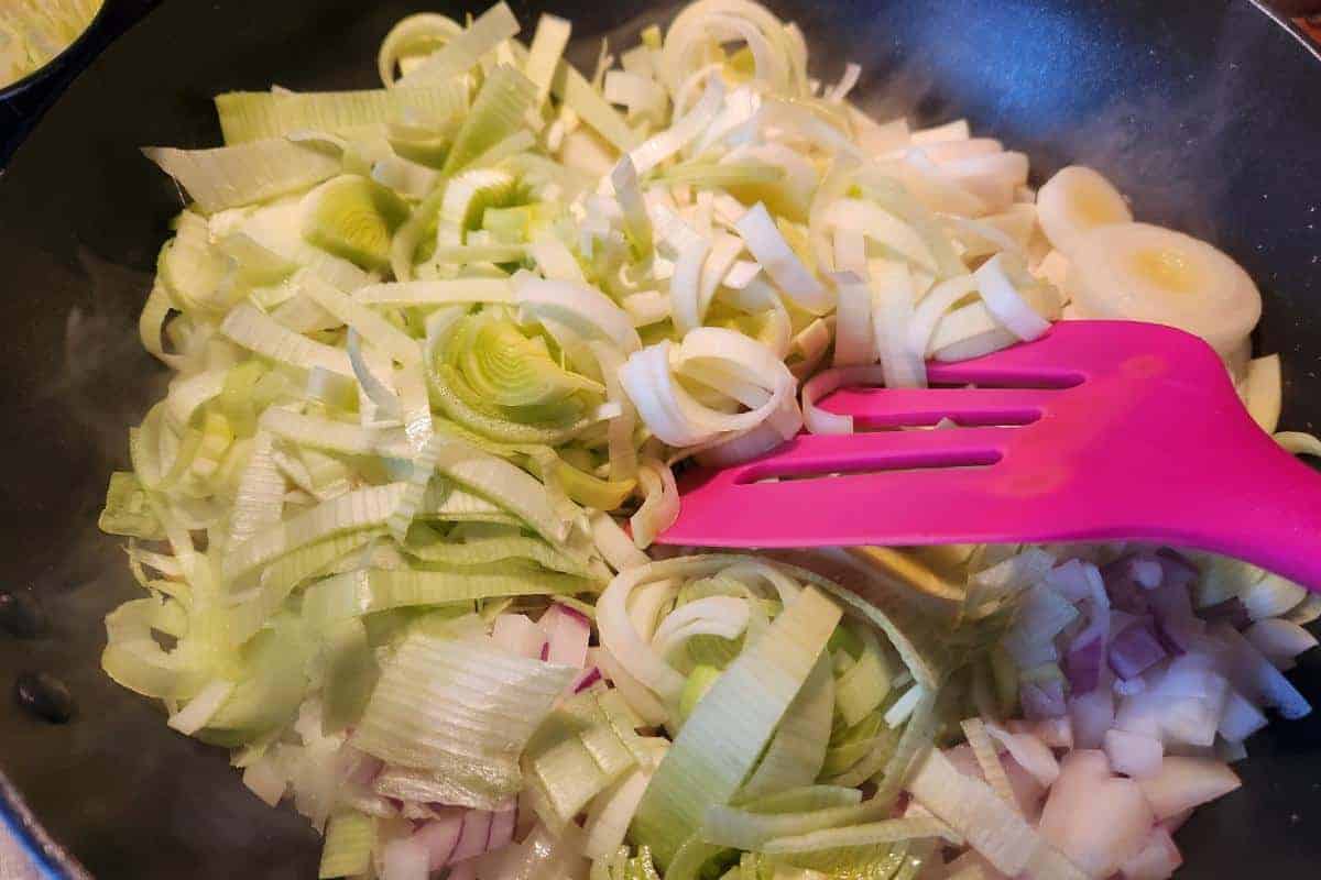 A frying pan containing red onion and sliced leeks with a spatula stirring them.