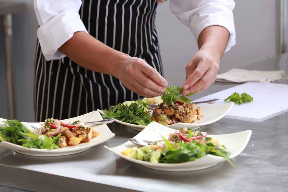 A chef standing preparing food of salad and accompaniments on square plates.
