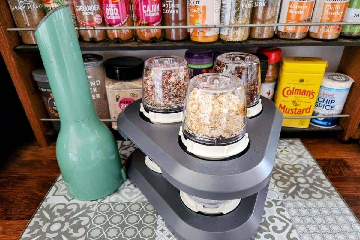 A Finamill spice grinder machine with four containers of assorted herbs and spices rests on a patterned surface, accompanied by various spice jars and a vibrant green electric pepper grinder.
