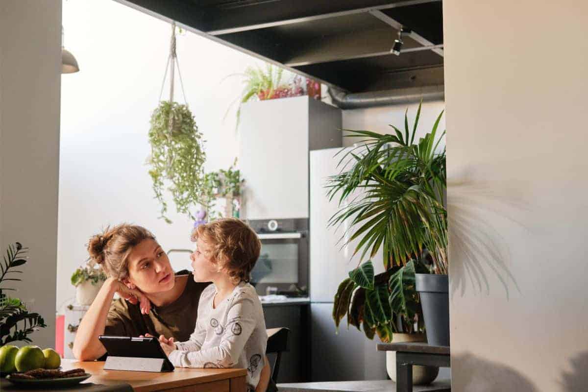 A woman and child sit at a table in a plant-filled kitchen. The woman listens attentively as the child, engaged in speech therapy activities, holds a tablet.
