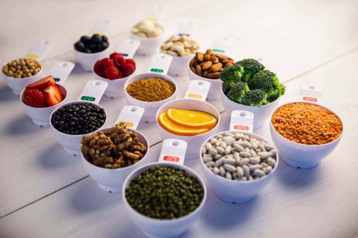 Measuring cups, ideal for portion control, are filled with an array of foods: vibrant berries, crunchy nuts, fresh broccoli, zesty citrus slices, hearty beans, nutritious lentils, pungent garlic, and healthy seeds—all beautifully arranged on a pristine white surface.