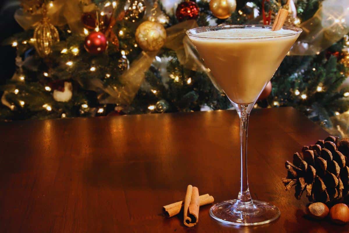 Christmas cocktails are tasty and this one is a cocktail glass with cinnamon sticks and eggnog with a Christmas tree in the background.