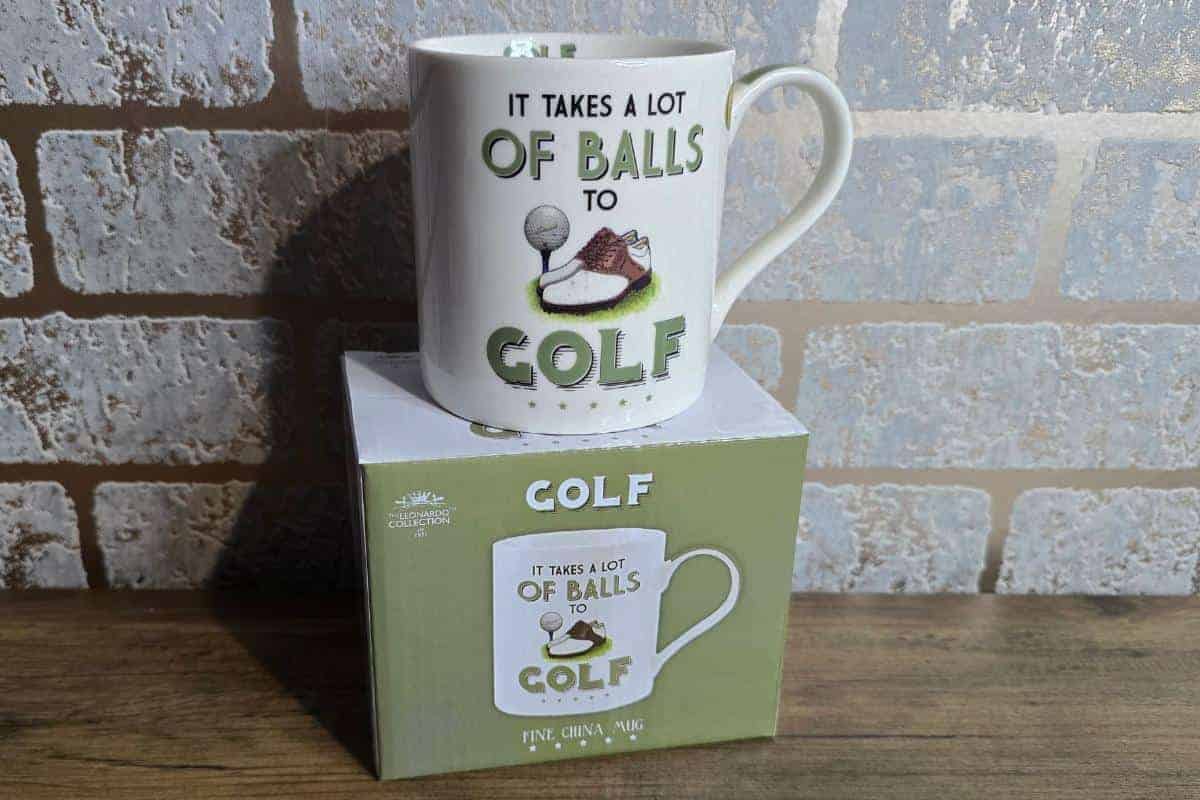 A ceramic golf-themed mug, perfect for men, sits on a matching box featuring the text "IT TAKES A LOT OF BALLS TO GOLF" along with a golf ball and shoes. This Cotton Traders item is among the top gift ideas for enthusiasts of the sport.