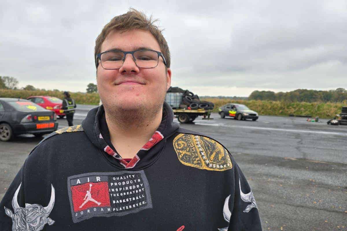 A person in a patterned hoodie stands on a road with several parked cars in the background under a cloudy sky, reviewing their thrilling passenger drifting experience.