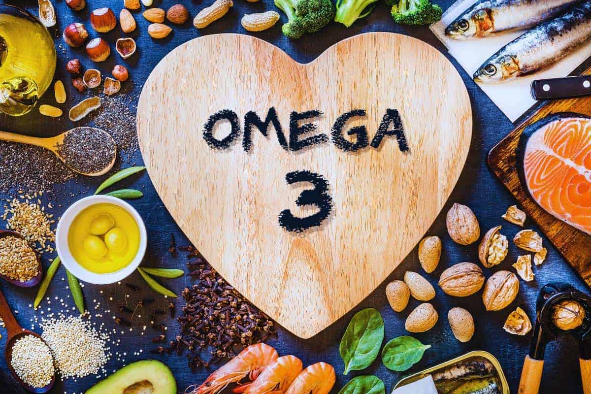 Heart-shaped board with "Omega 3" text, surrounded by salmon, sardines, nuts, seeds, avocado, broccoli, and shrimp—showcasing the benefits of these nutrient-rich foods. Nearby are bowls of oil and eggs on a dark surface.