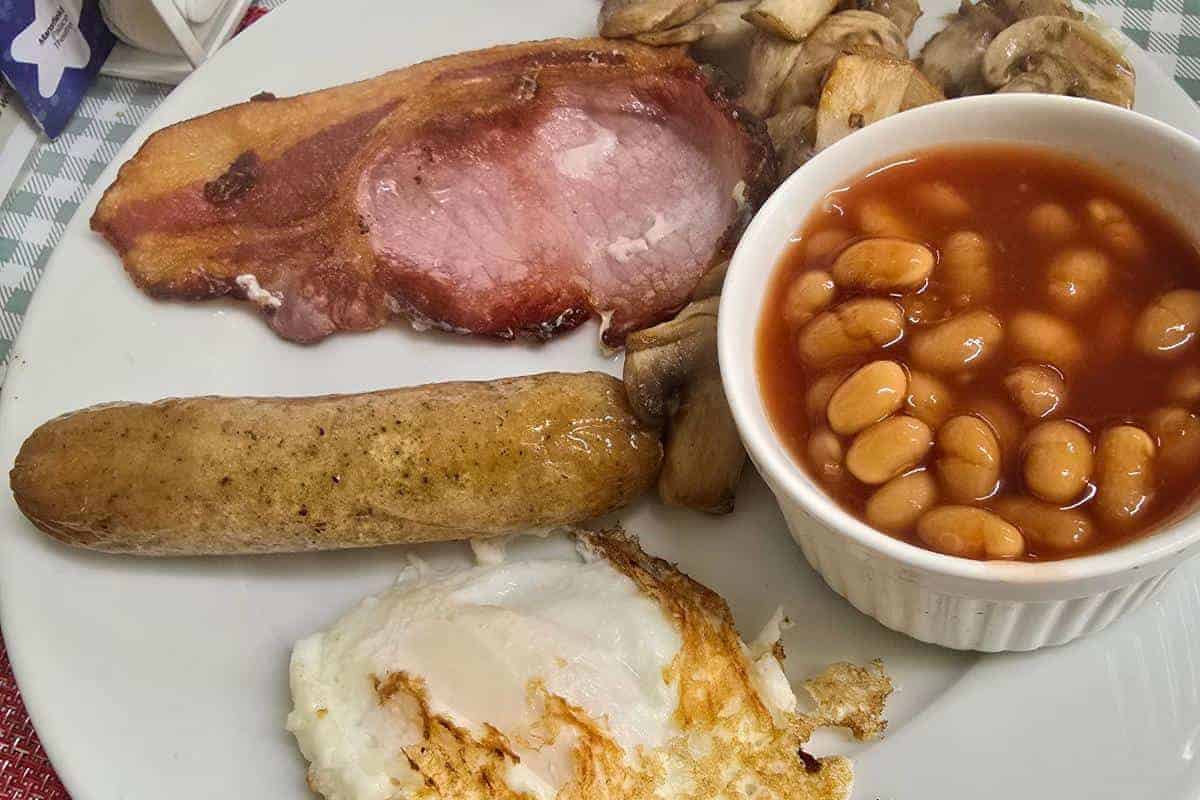 Plate with a fried egg, sausage, slice of bacon, mushrooms, and a small bowl of baked beans—your breakfast sweet spot. Perfectly arranged at PoFfles Coffee and Cake in Mansfield.