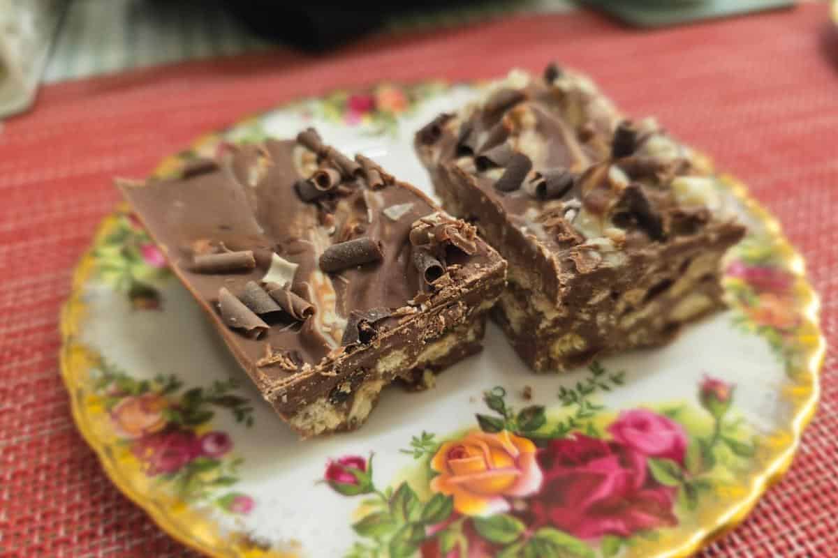 Two chocolate squares on a decorative floral plate, topped with chocolate curls and a nutty layer, set on a red placemat—perfectly paired with coffee from Sweet Spot Mansfield.