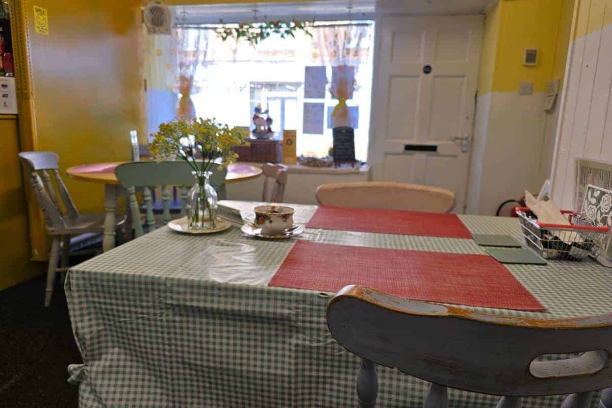 The small cafe, known for its delightful PoFfles, features a cozy interior with green checkered tablecloths, red placemats, and a vase of flowers on each table. The yellow walls and window offering an inviting view outside enhance the welcoming atmosphere.