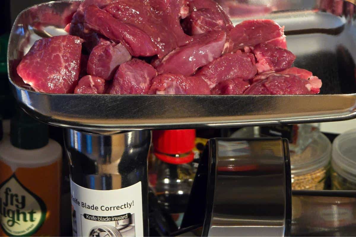 Chunks of raw red meat sit on a metal tray above an AAOBOSI Electric Meat Grinder, 3000W, with various kitchen items in the background.