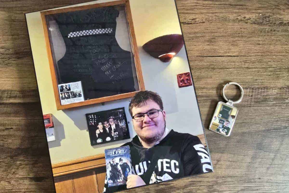 A person holding a "Fast & Furious" DVD stands in front of a framed black vest on the wall, perhaps contemplating an unconventional Mother's Day gift. A keychain with a similar image sits on the wooden table beside them.