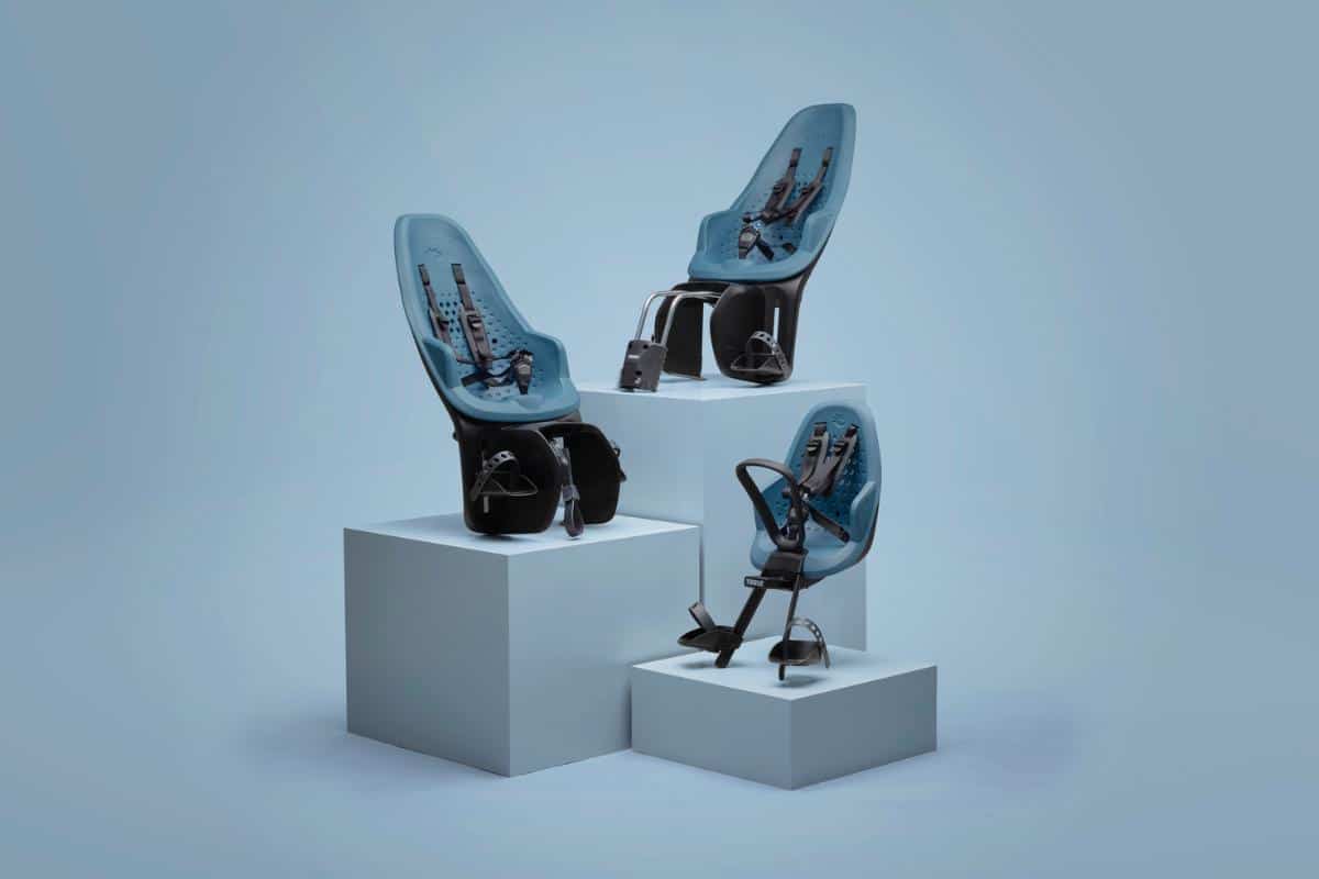 Three transparent blue child car seats with visible internal harnesses are displayed on white cubic pedestals against a light blue background. One seat is smaller and placed in front of the others.