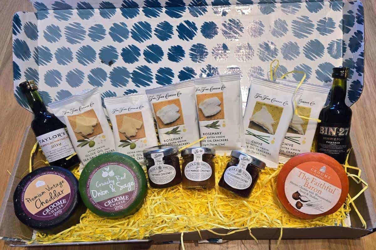 A gift box filled with yellow shredded paper contains two bottles of port, cheese rounds, six packs of assorted cheese crackers, and three small jars of preserves or chutneys, all neatly arranged inside a blue and white patterned box.