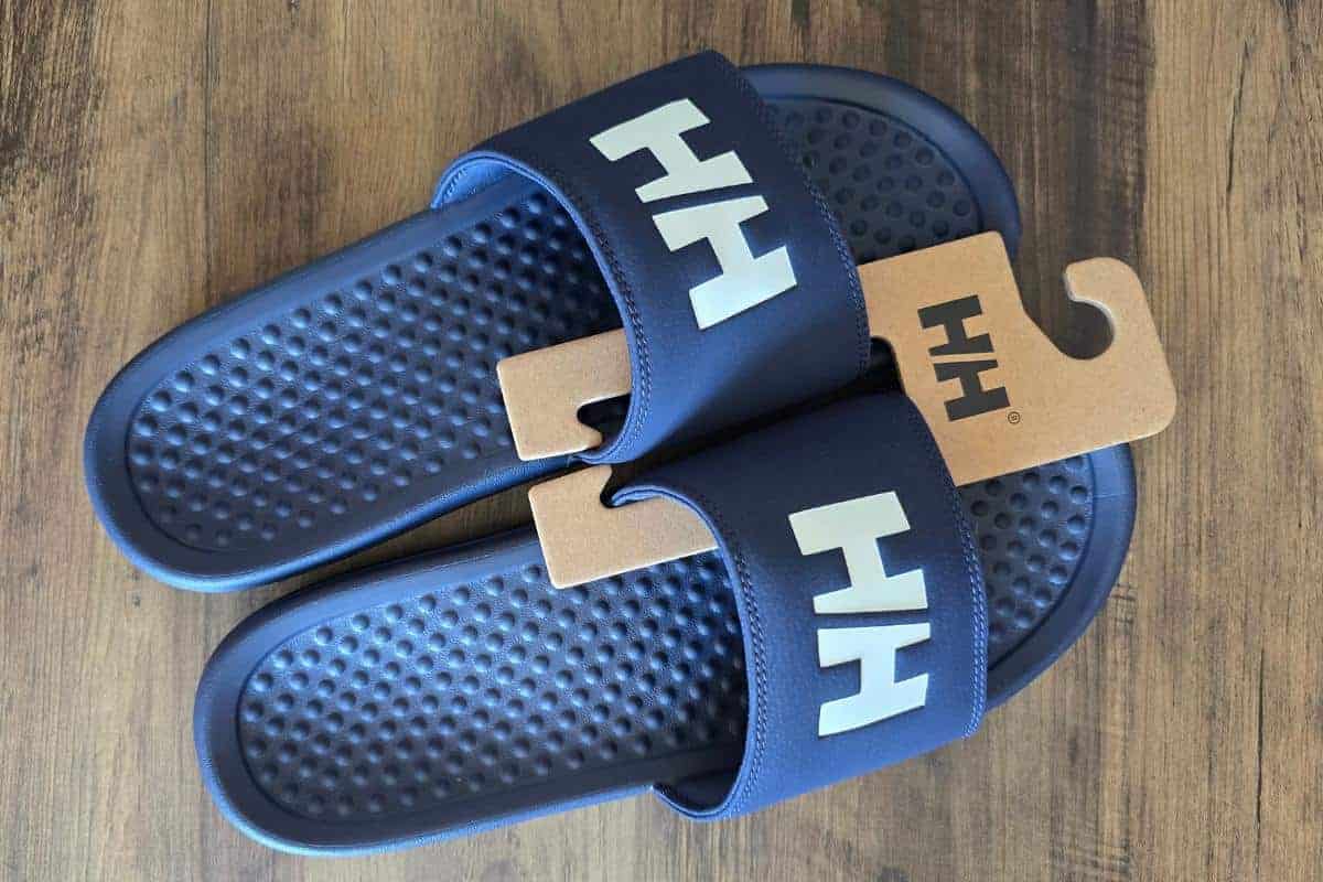 A pair of navy blue HH brand slide sandals with textured footbeds, displayed overlapping on a wooden surface. They are held together by a brown cardboard hanger. The white "HH" logo is prominently printed on each sandal’s strap.