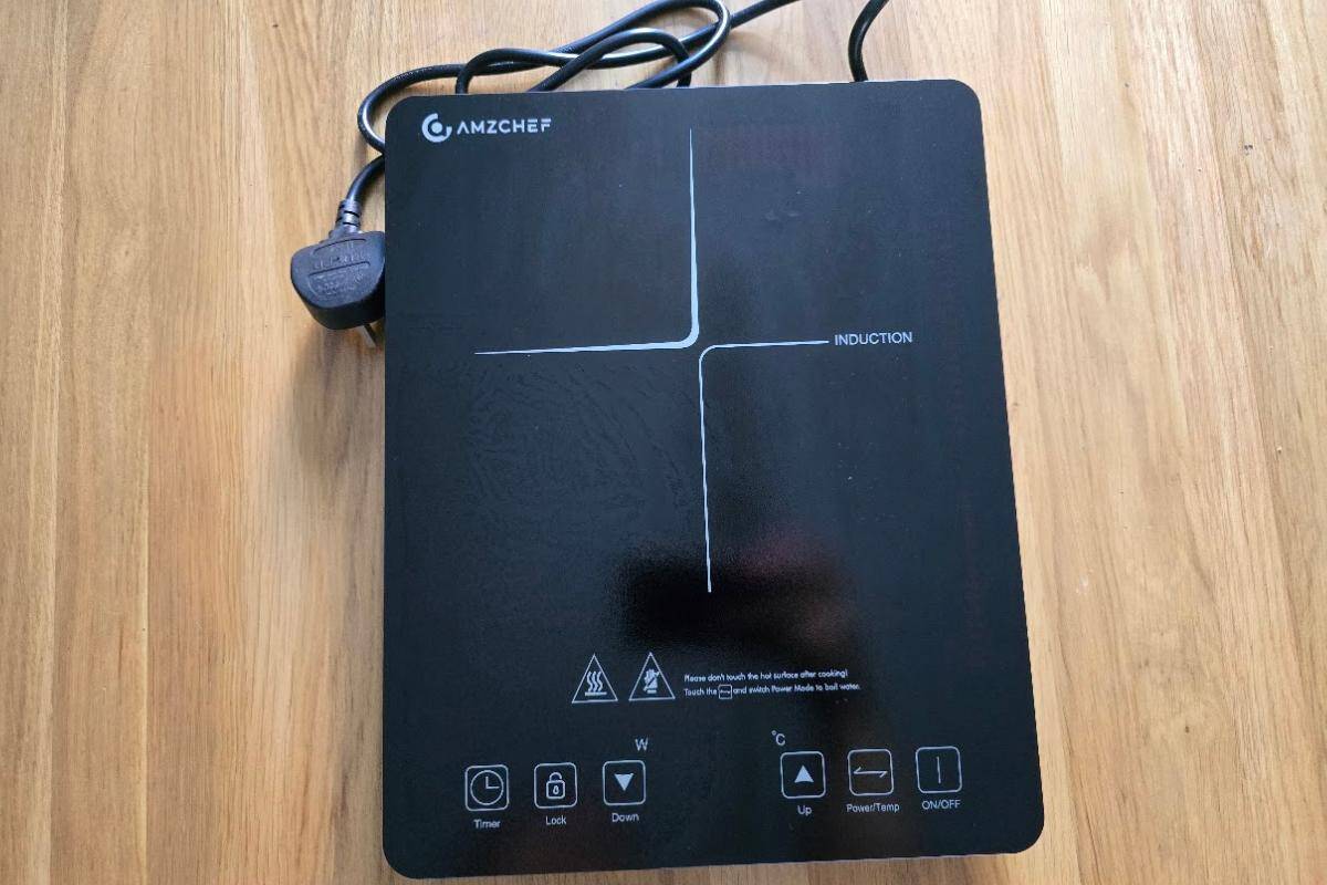 A black AMZCHEF single induction hob with a touch control panel sits on a wooden surface. Showcasing compact convenience, the power cord and plug are visible at the top. The cooktop features timer, lock, temperature, and power buttons.
